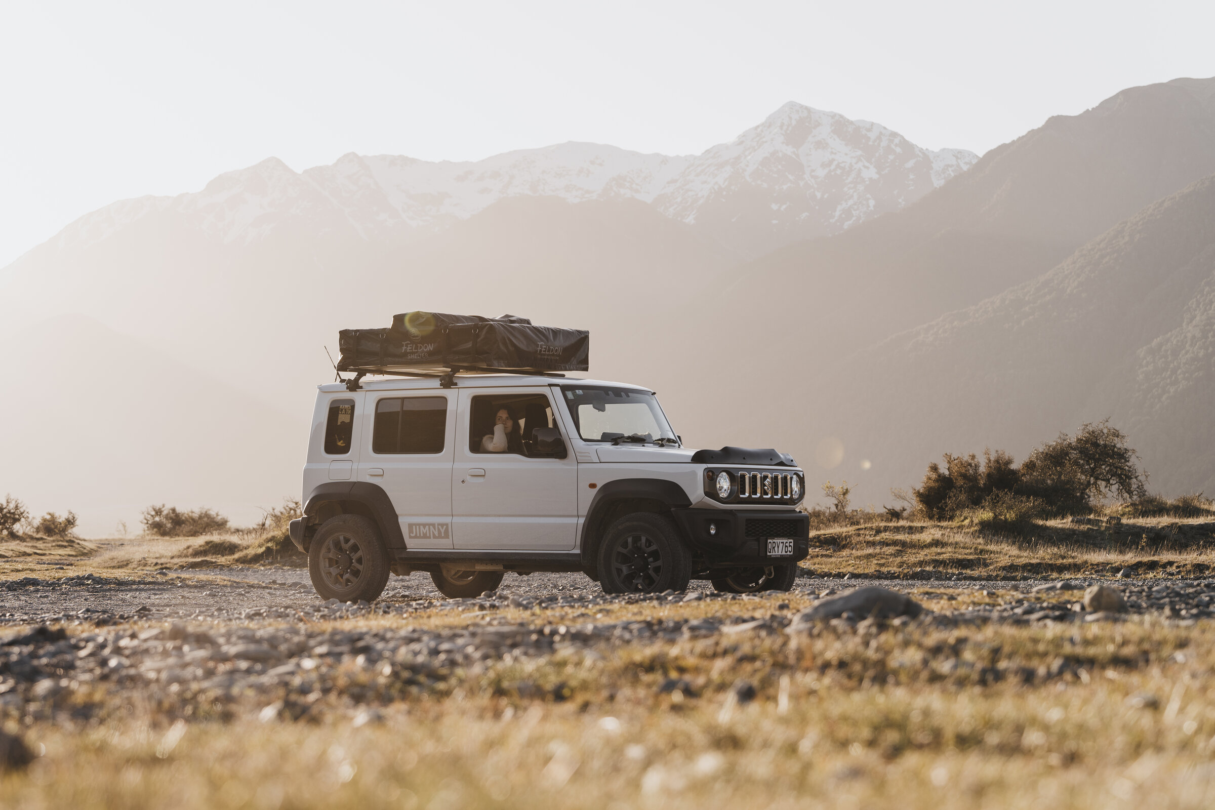 White 5-door Suzuki Jimny with rooftop tents at golden hour, snow-capped South Island mountains behind