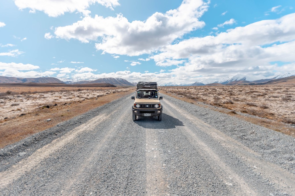 Tan Suzuki Jimny with rooftop tent on gravel road with snow-capped mountains, South Island.