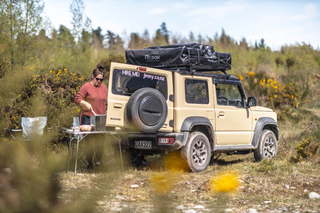 White Suzuki Jimny with rooftop tent in golden hour, mountain backdrop.
