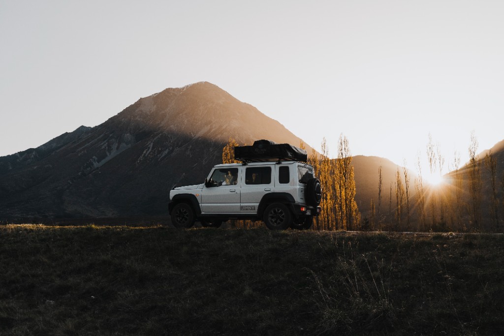 Jimny parked by water with mountain backdrop.