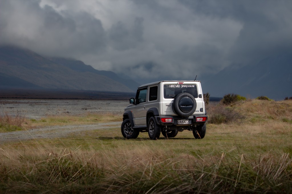 Woman beside white Jimny with deployed rooftop tent and ladder, lakeside.