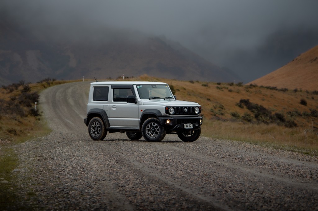 Tan Suzuki Jimny with rooftop tent driving on gravel road, plains and snow-capped mountains.