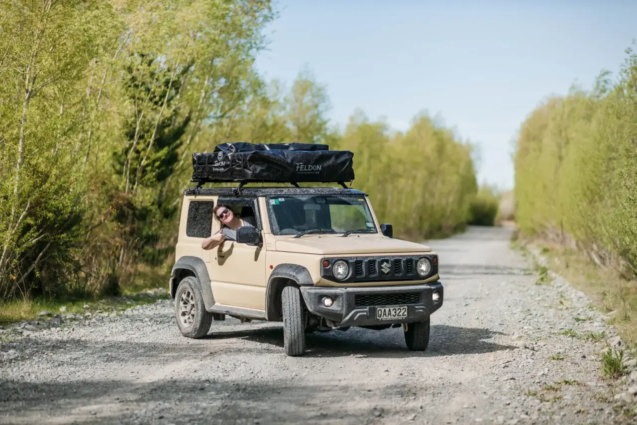White Suzuki Jimny with two Feldon Shelter rooftop tents on gravel road, mountain vista. – Jimny Rentals.