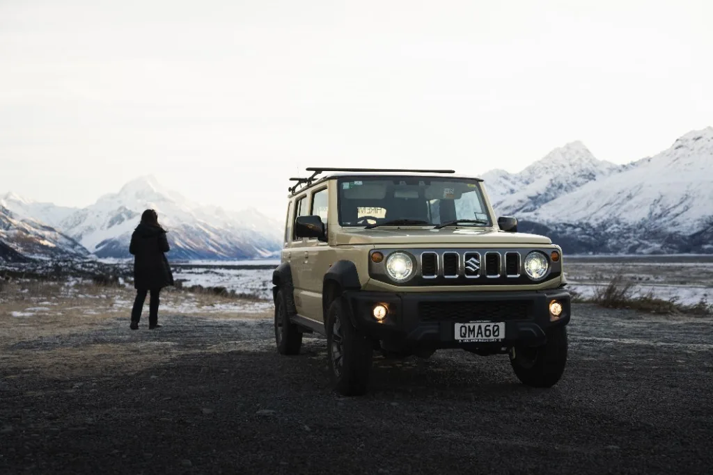 Tan Suzuki Jimny on a gravel road with snow-capped South Island mountains and a person enjoying the view.