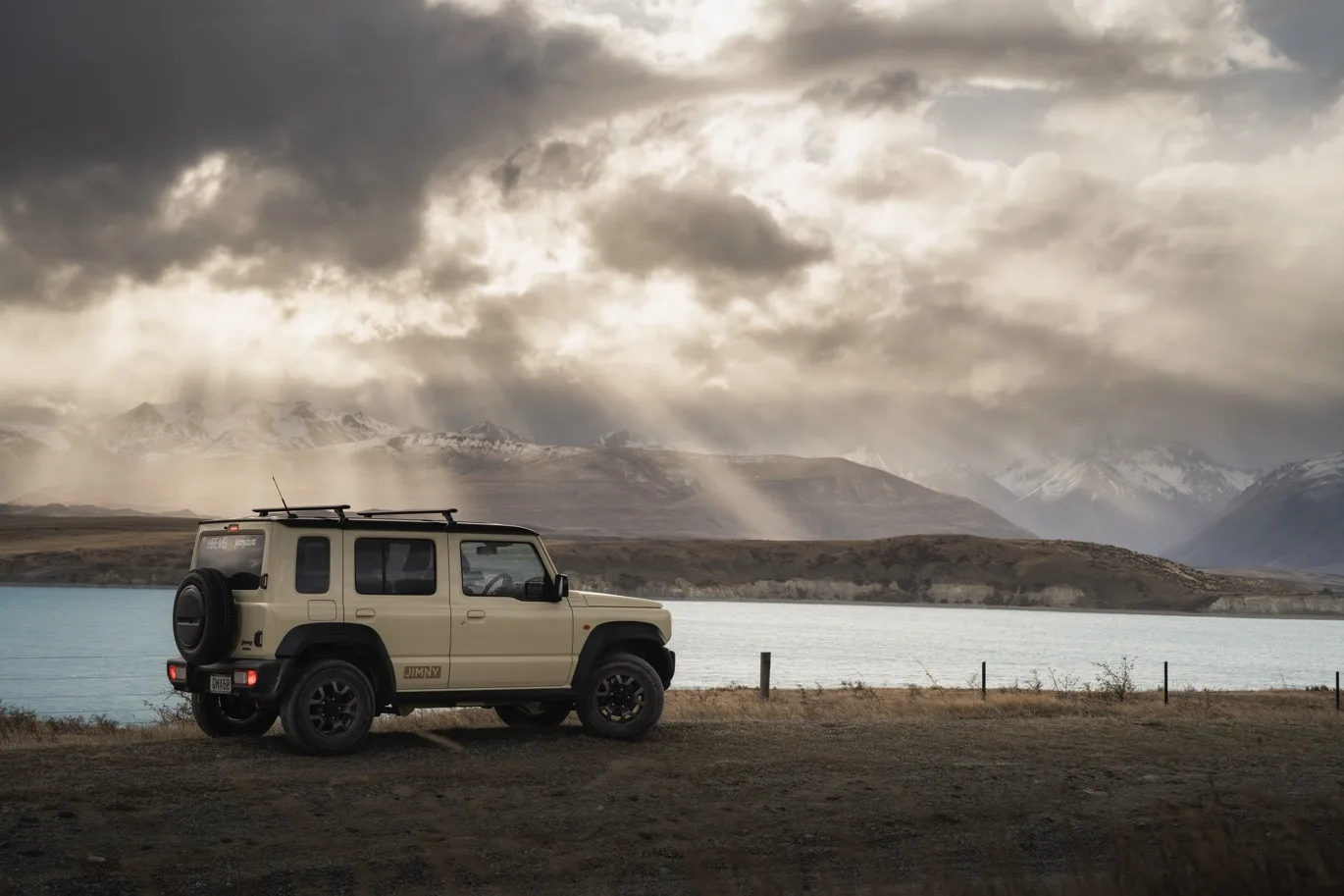 Suzuki Jimny driving on winding road with snow-capped mountains. – Jimny Rentals.