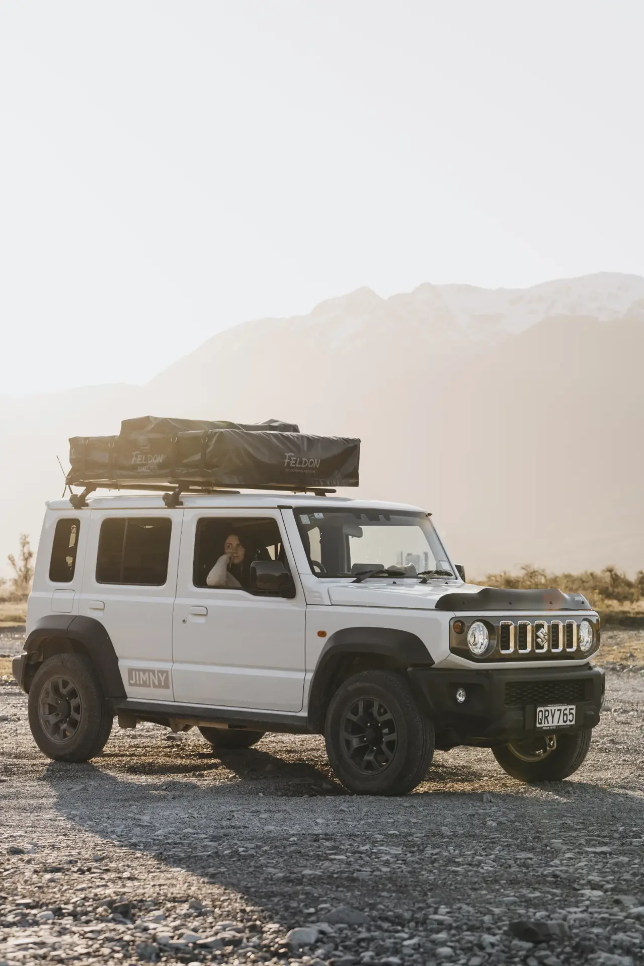 White Suzuki Jimny with rooftop tent in grassy field, mountains behind. – Jimny Rentals.