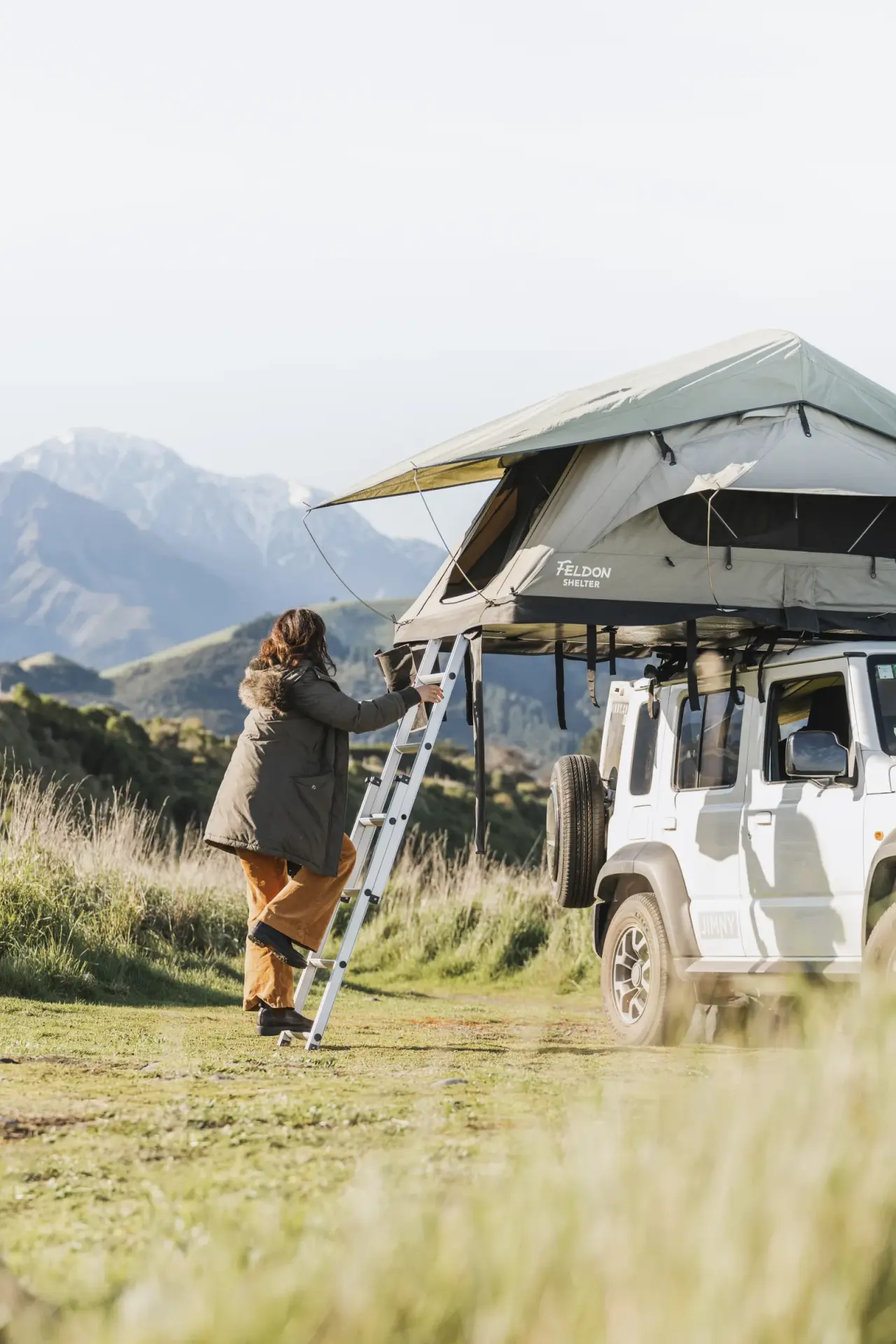 White Suzuki Jimny with rooftop tent on gravel track, snow-capped mountains. – Jimny Rentals.