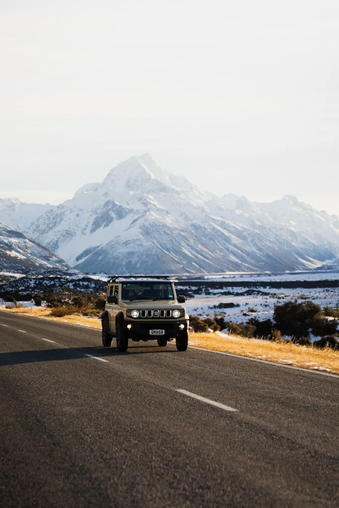 Tan Suzuki Jimny on a paved road with a large snow-capped mountain behind, golden hour.
