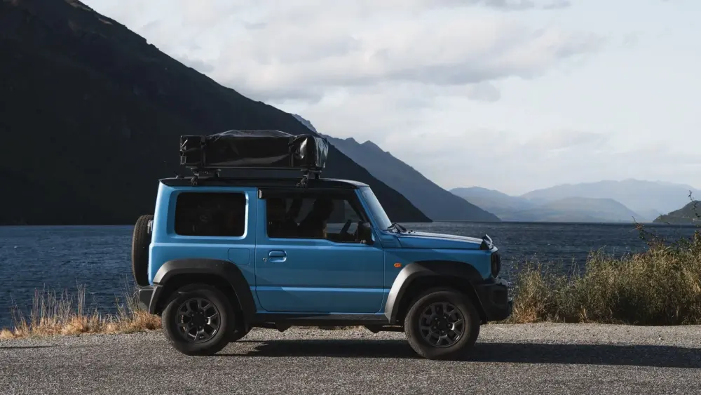 A Jimny parked by a South Island lake edge with mountains behind