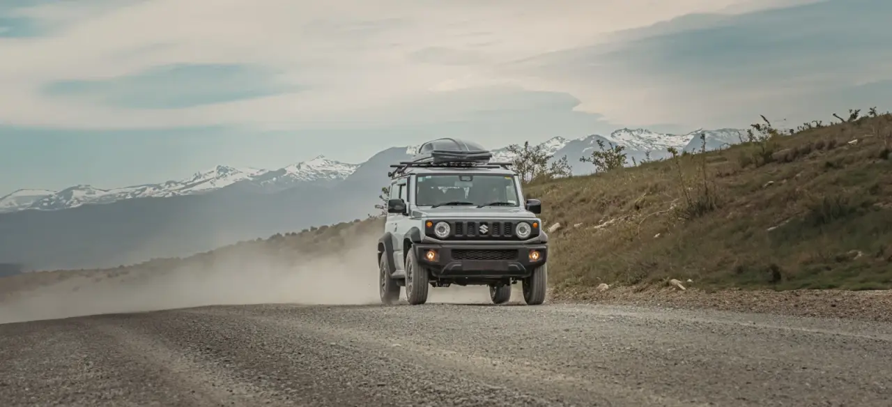 White Suzuki Jimny with two rooftop tents on gravel road, driver at window, mountain backdrop. – Jimny Rentals.